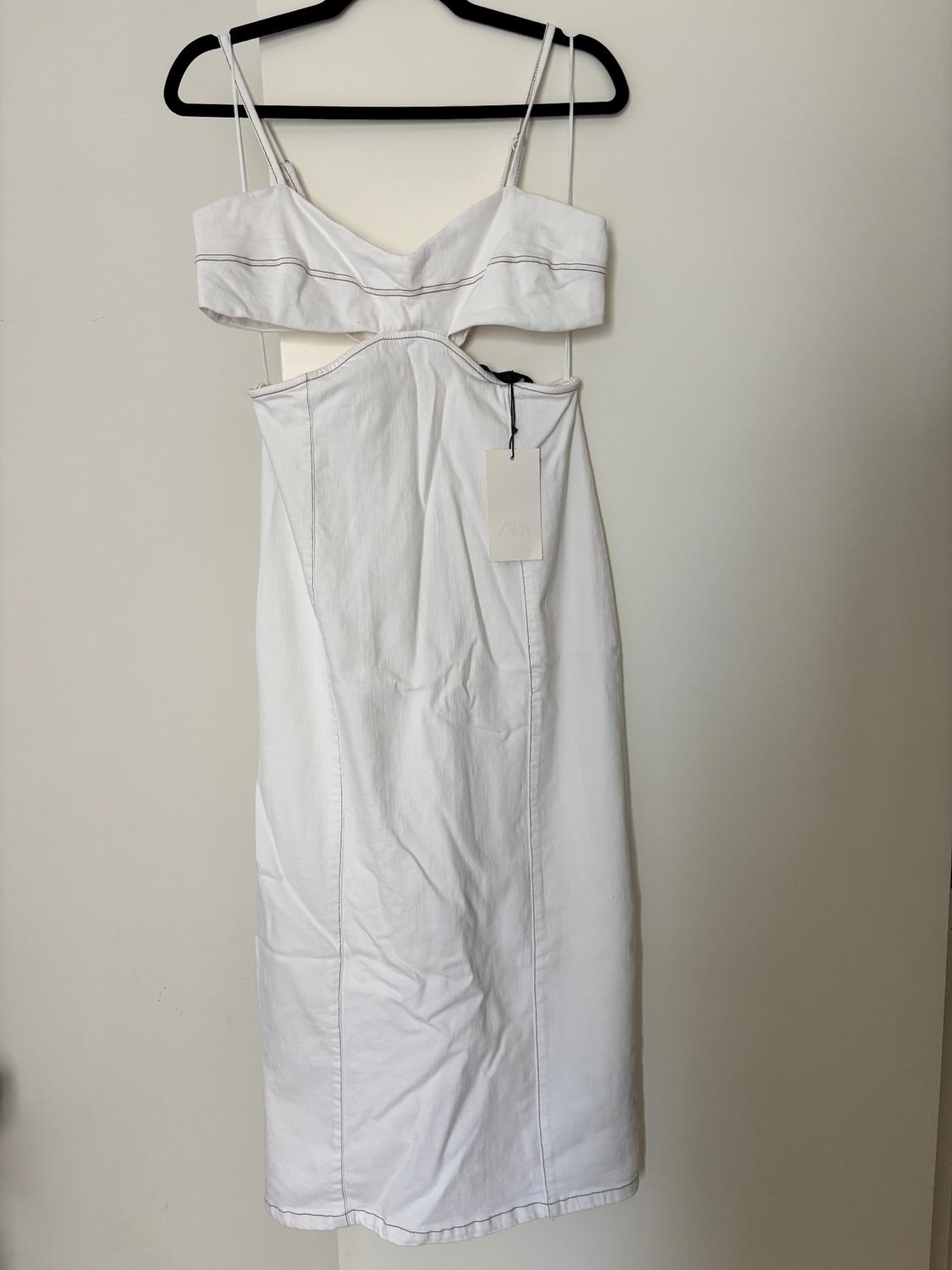 White dress with spaghetti straps and a cut-out waist, hanging on a black hanger. Label visible in the centre of the dress.