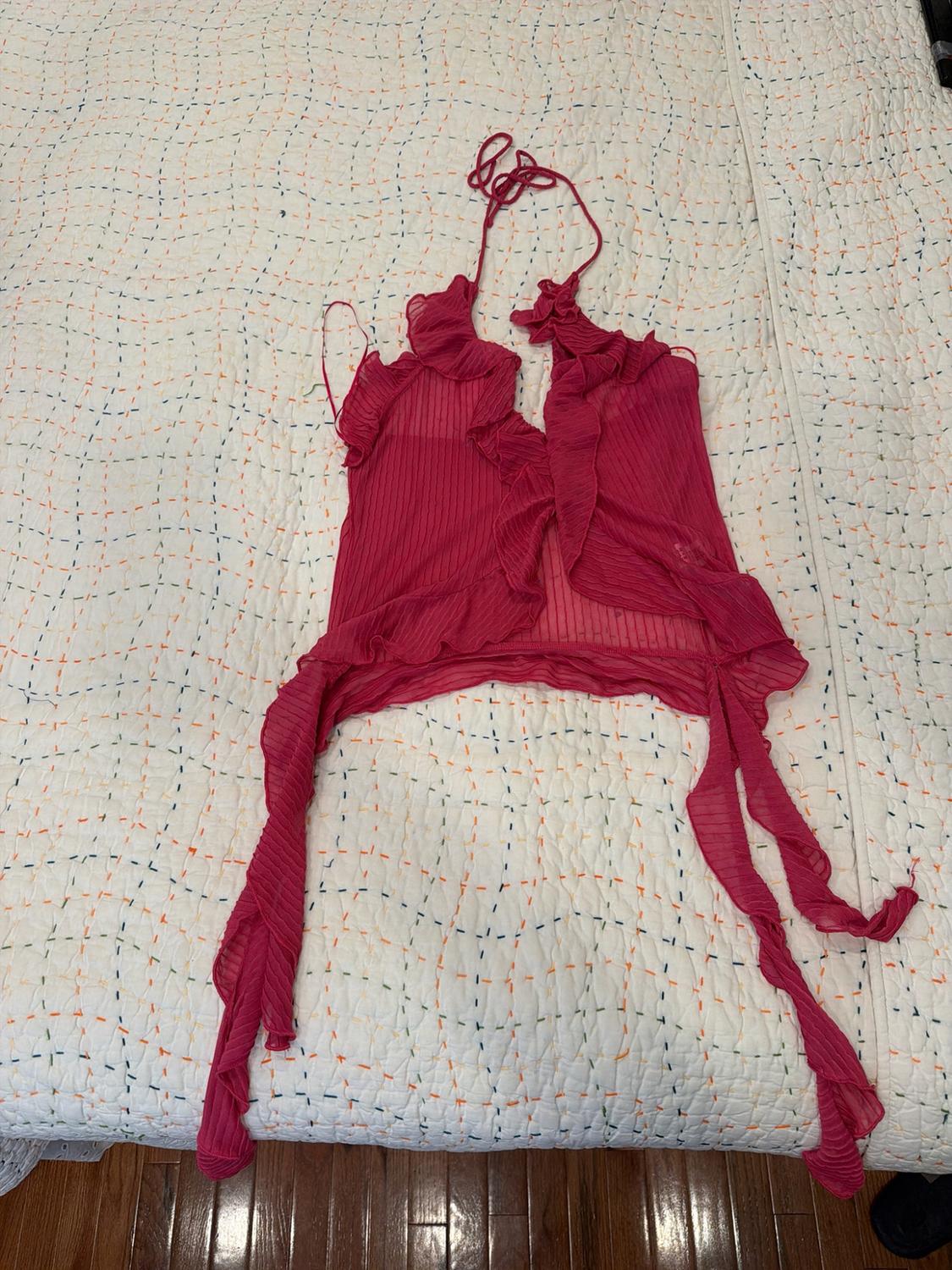 Sheer fuchsia halter top with ruffles and long straps, on a white bedspread with colorful details. Visible wooden floor.