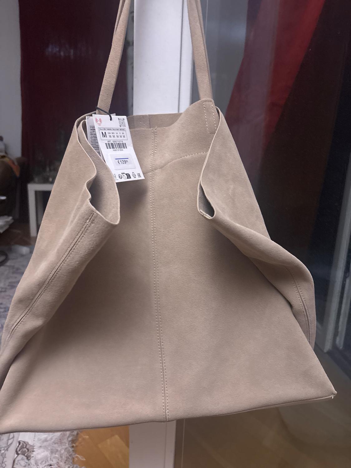 Hanging beige suede tote bag. Shows a label with the text “M” and “€129.00”. Interior of a house in the background with a red curtain.