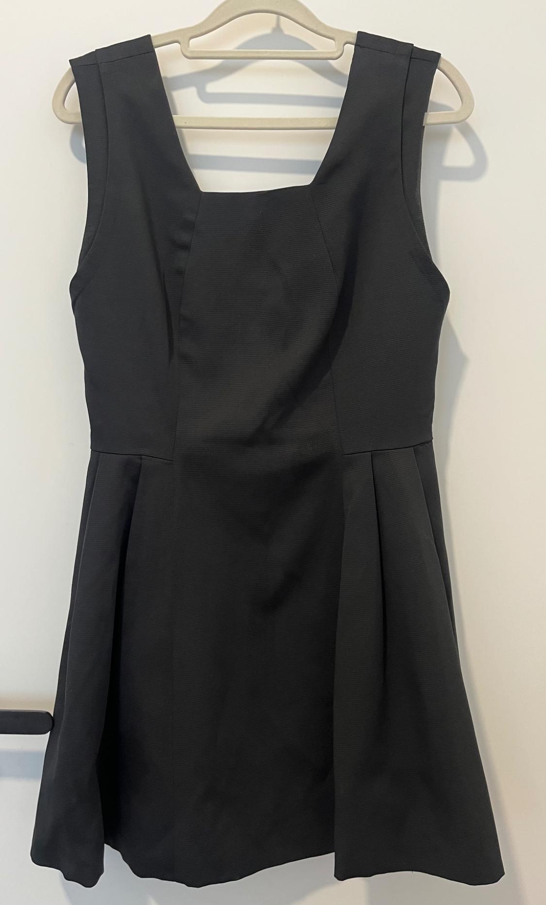 Sleeveless black dress hanging on a beige hanger, with a square neckline and pleated skirt, on a white background.