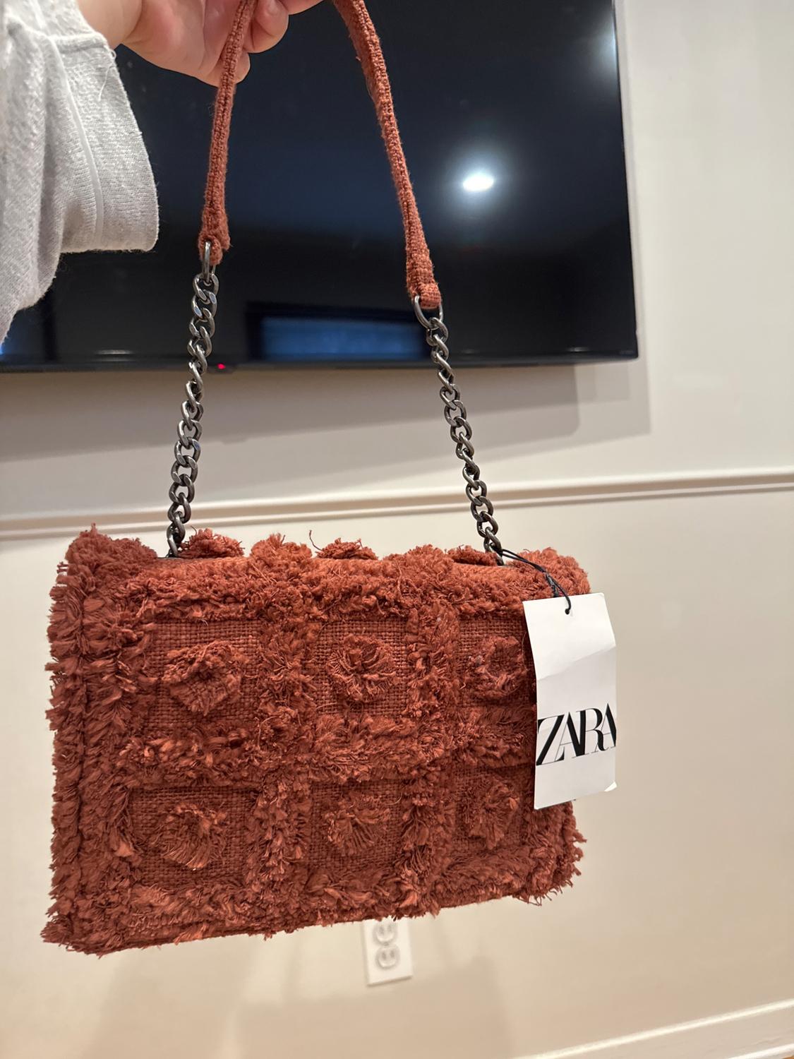Brown handbag with a furry texture and metal chain. Visible label with "ZARA" text. Interior background with white wall and television.