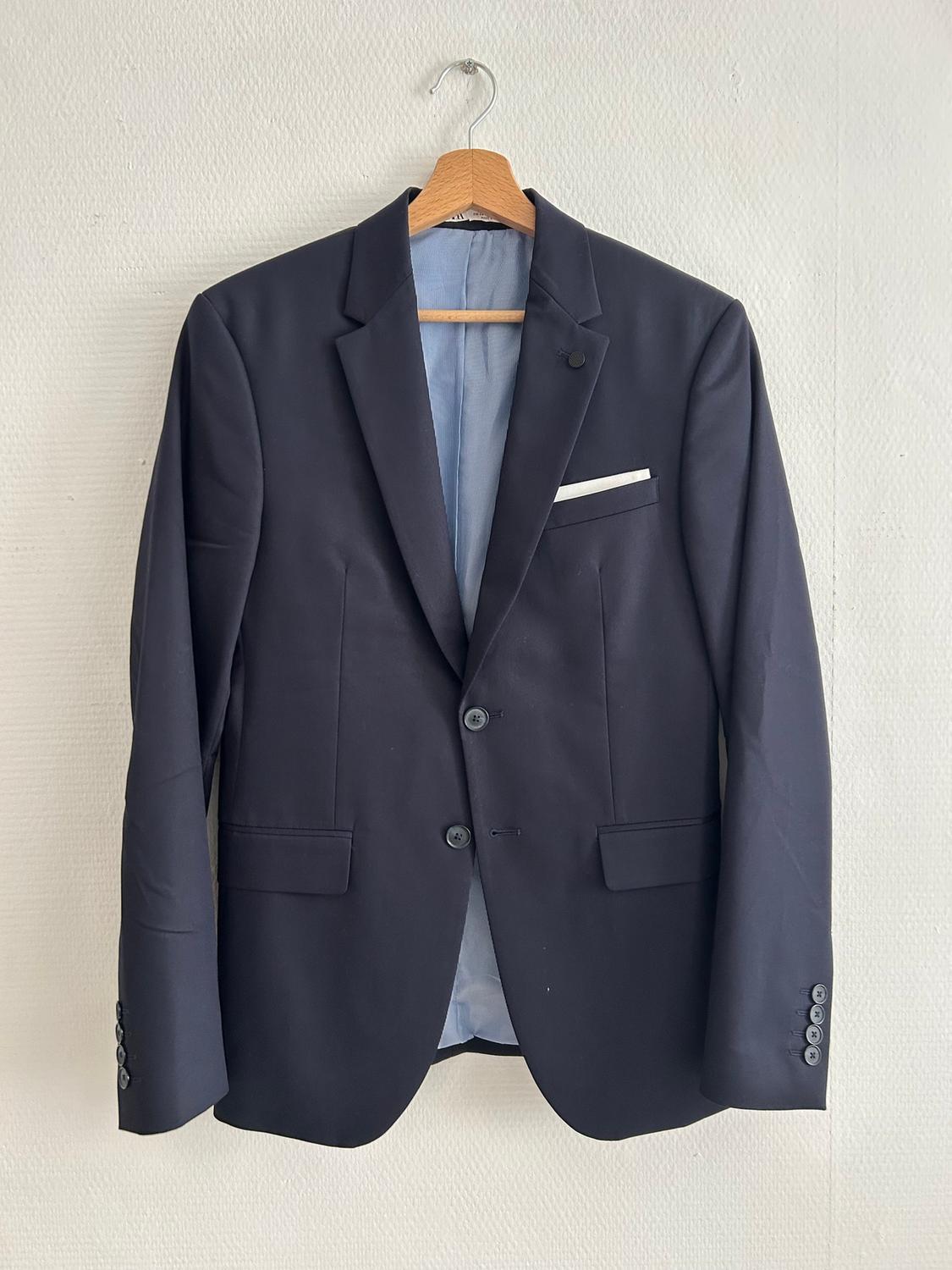 Black jacket hanging on a wooden hanger, with a classic lapel and a pocket with a white handkerchief, against a white wall background.