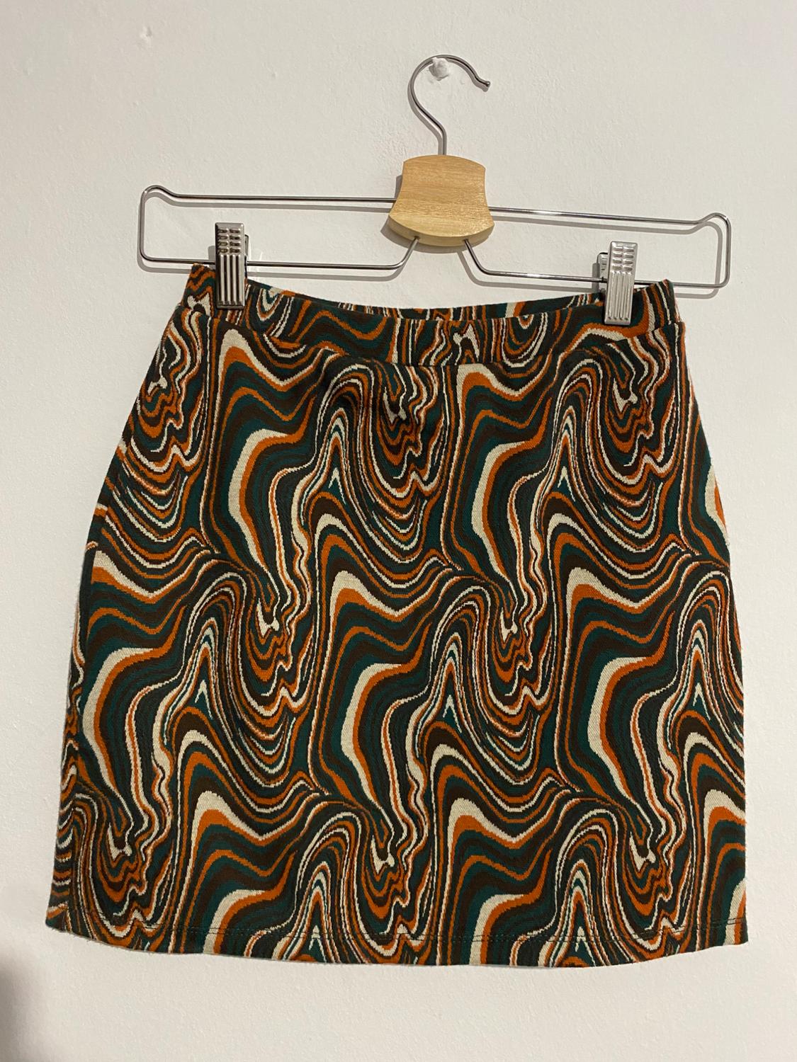 Short skirt with psychedelic print in brown, green and beige tones, hanging on a wooden hanger against a white background.