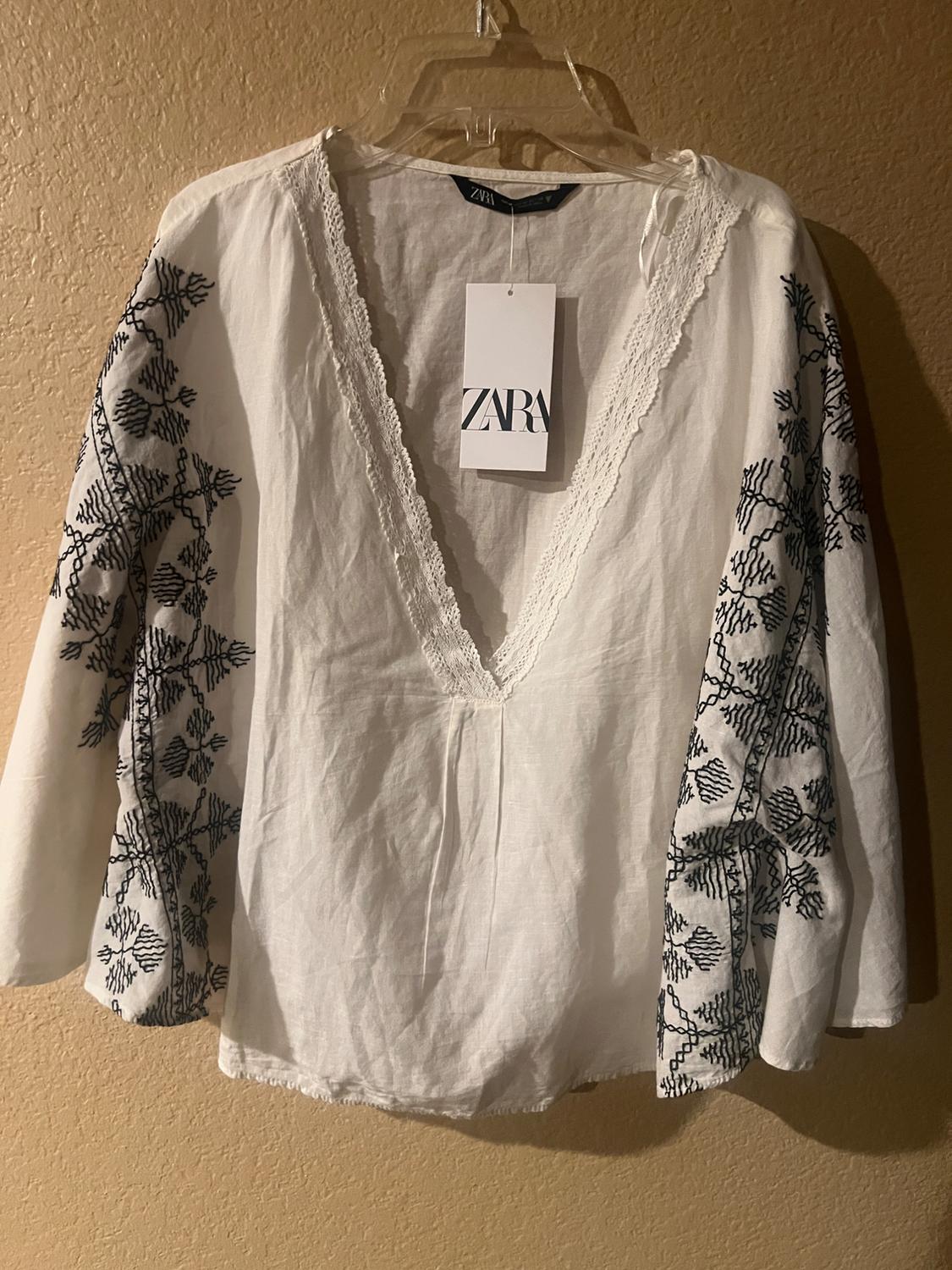 White blouse with black embroidery on the sleeves, V-neck and Zara tag hanging, on a transparent hanger.