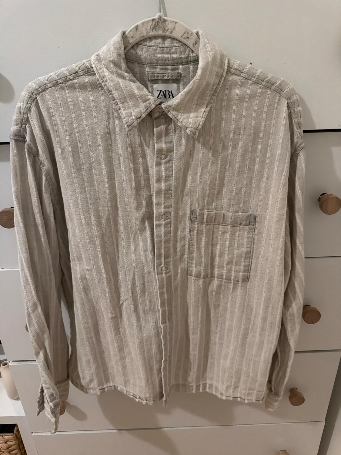 Beige vertical striped shirt hanging on a hanger, with a chest pocket and a white chest of drawers background. Visible label: ZARA.