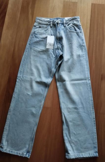 Light blue straight-cut jeans on a wooden floor, with a white hanging tag that says "ZARA".