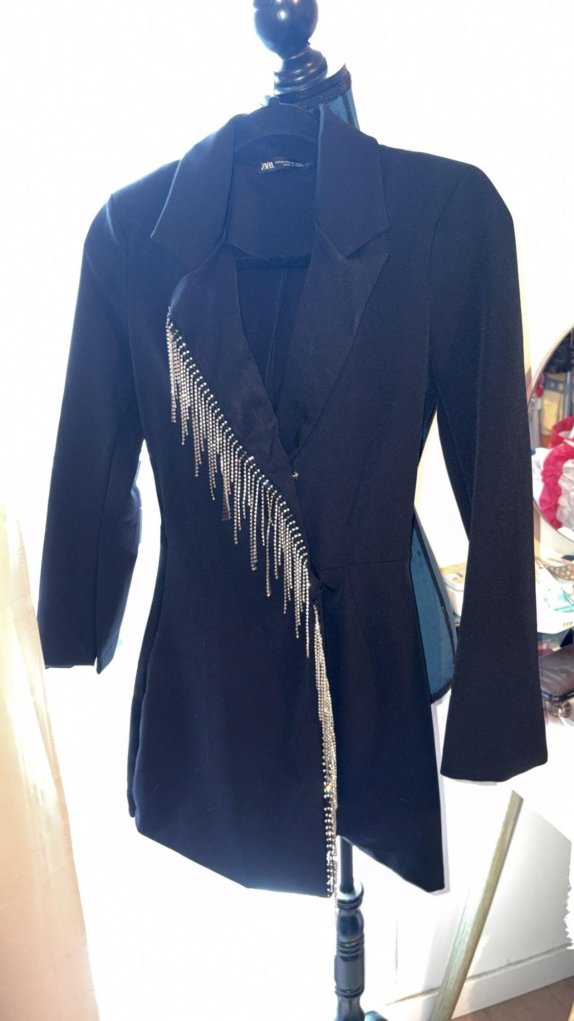 Μαύρο blazer με πέτα και διαγώνιες διακοσμήσεις από λαμπερές αλυσίδες, κρεμασμένο σε μια κούκλα πάνω σε ανοιχτόχρωμο φόντο.