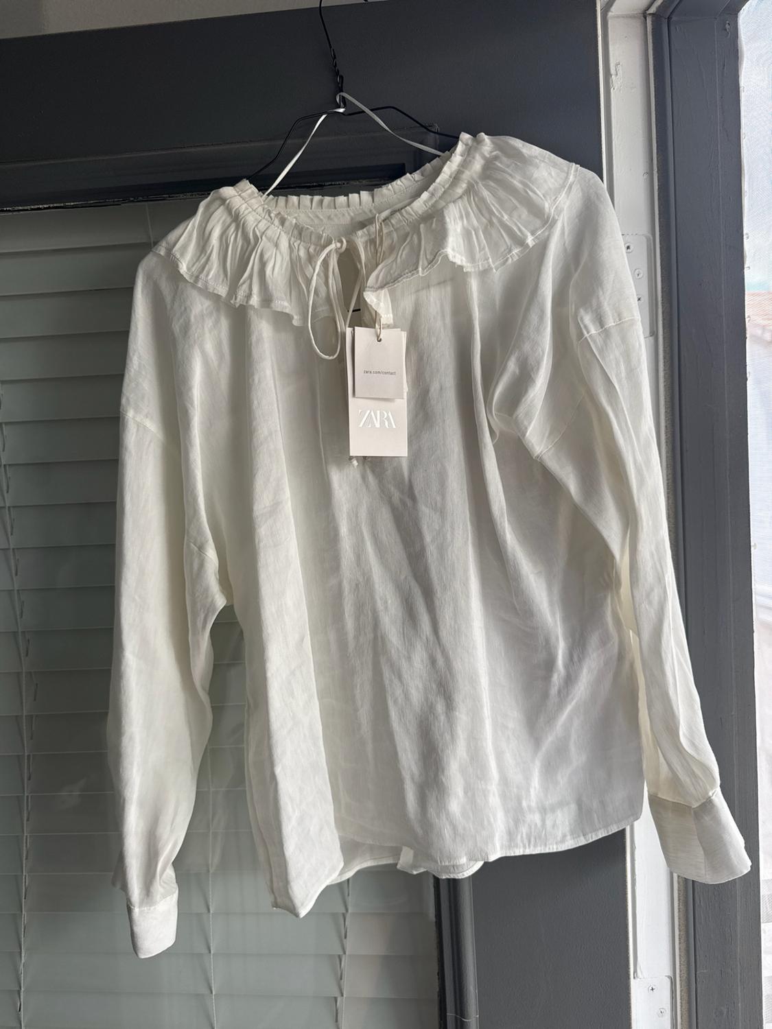 White long-sleeved blouse with a ruffled collar, hanging on a hanger in front of a window with blinds. Tag with the text ZARA.