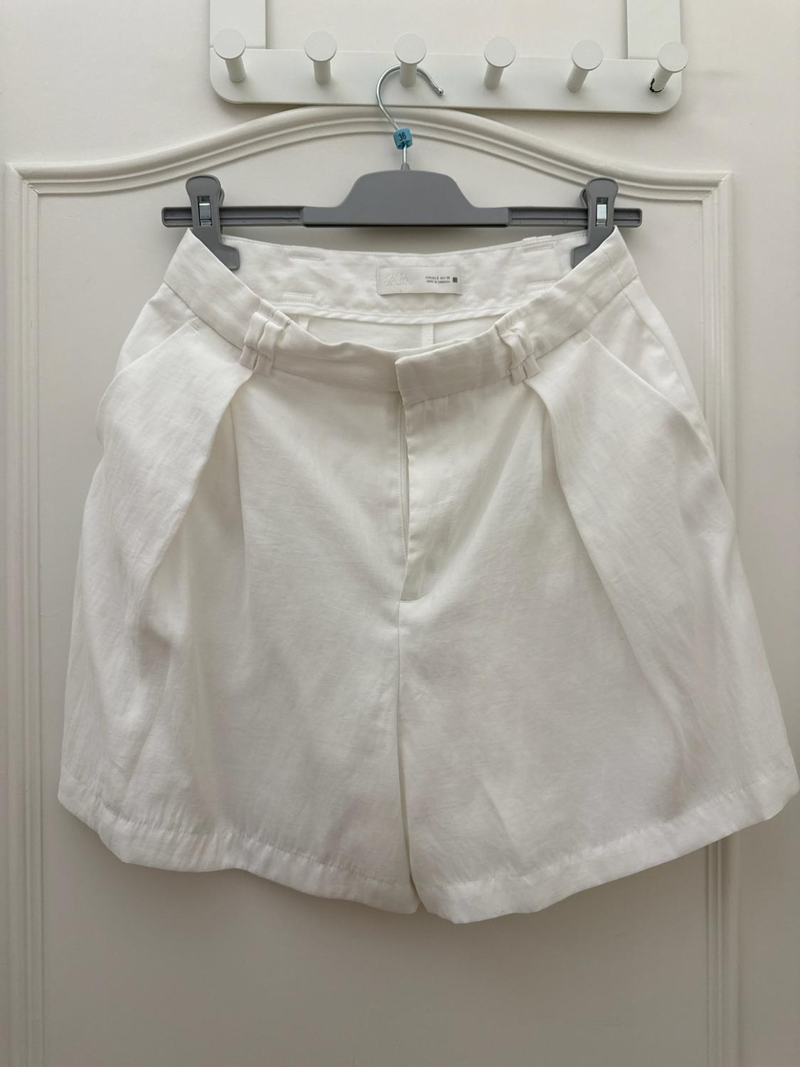 White tailored shorts hanging on a grey hanger on a white door. Inner label with the text ZARA.
