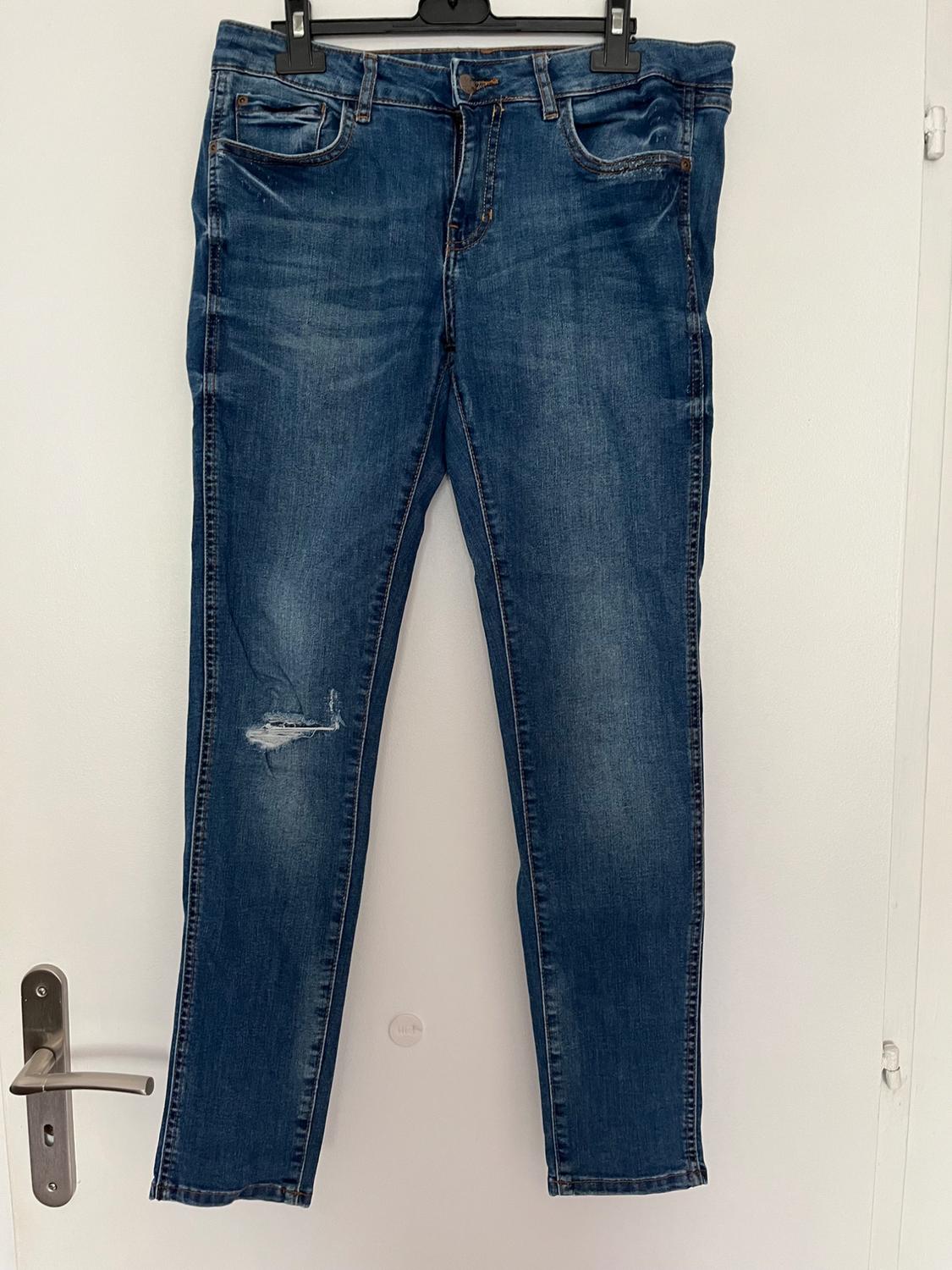 Front view of blue denim jeans with front pockets and distressed detail on the left leg.