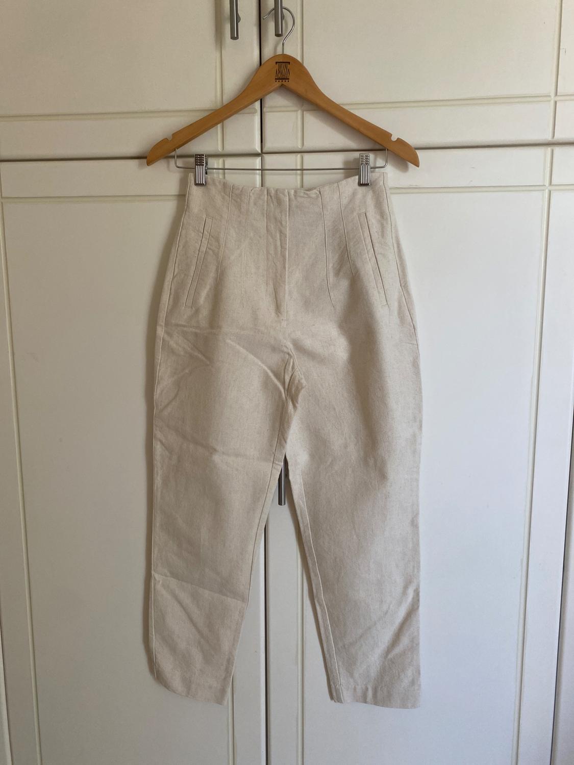 Light beige high-waisted trousers hanging in a white wardrobe on a wooden hanger that says BRIAN APOLLON. Casual and relaxed style.