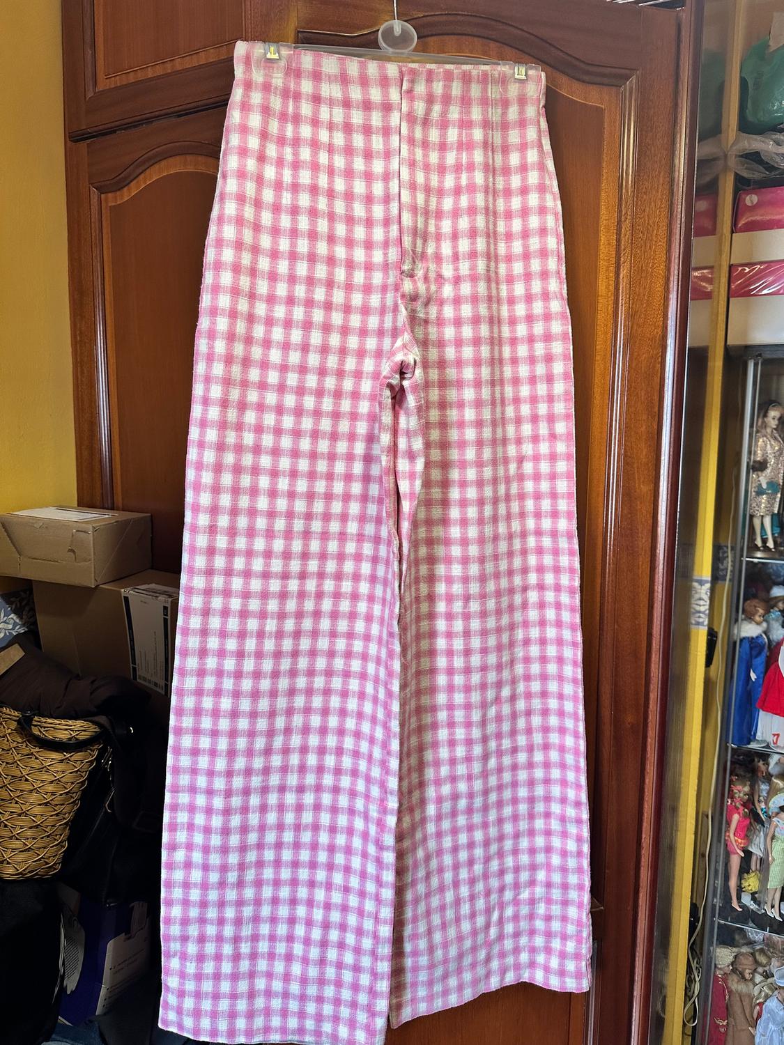 Pink and white checked trousers hanging on a wooden door. In the background, boxes and shelves with dolls.