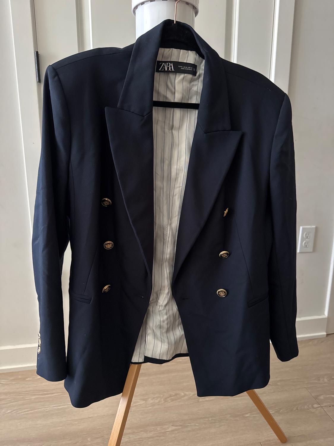 Black double-breasted jacket with gold buttons, hanging on a wooden coat rack. Interior with striped lining. Visible brand: Zara.