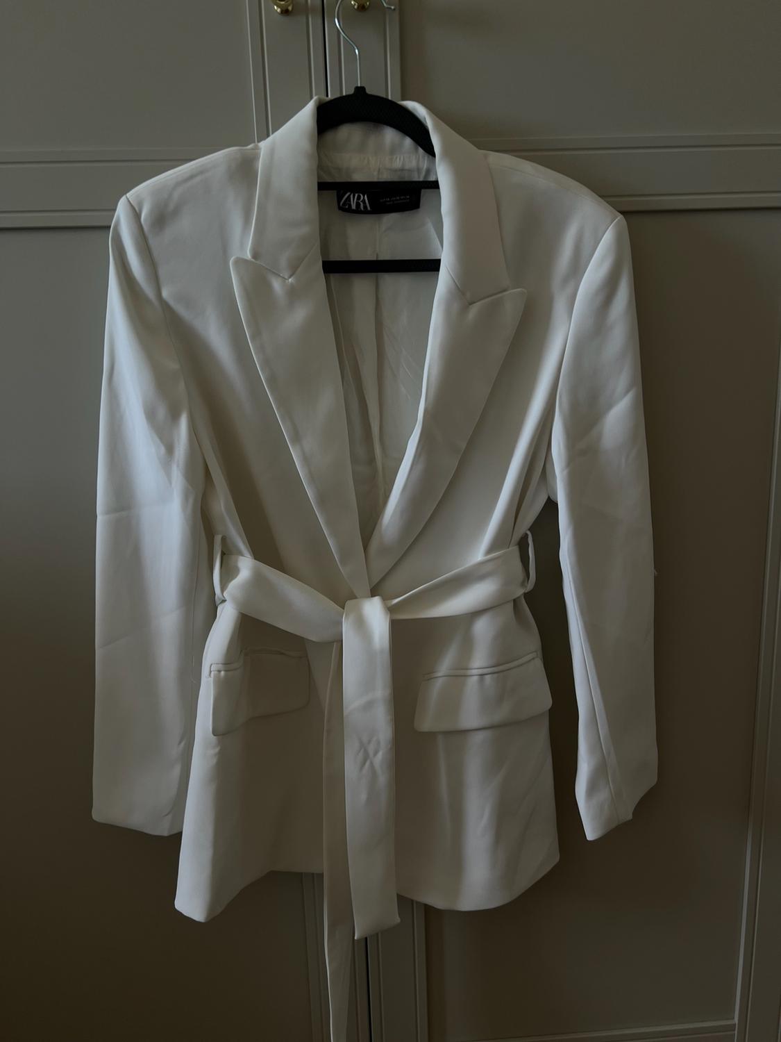 White blazer with lapels, pockets, and a tie belt, hanging on a black hanger in front of a wardrobe. Label with the text ZARA.