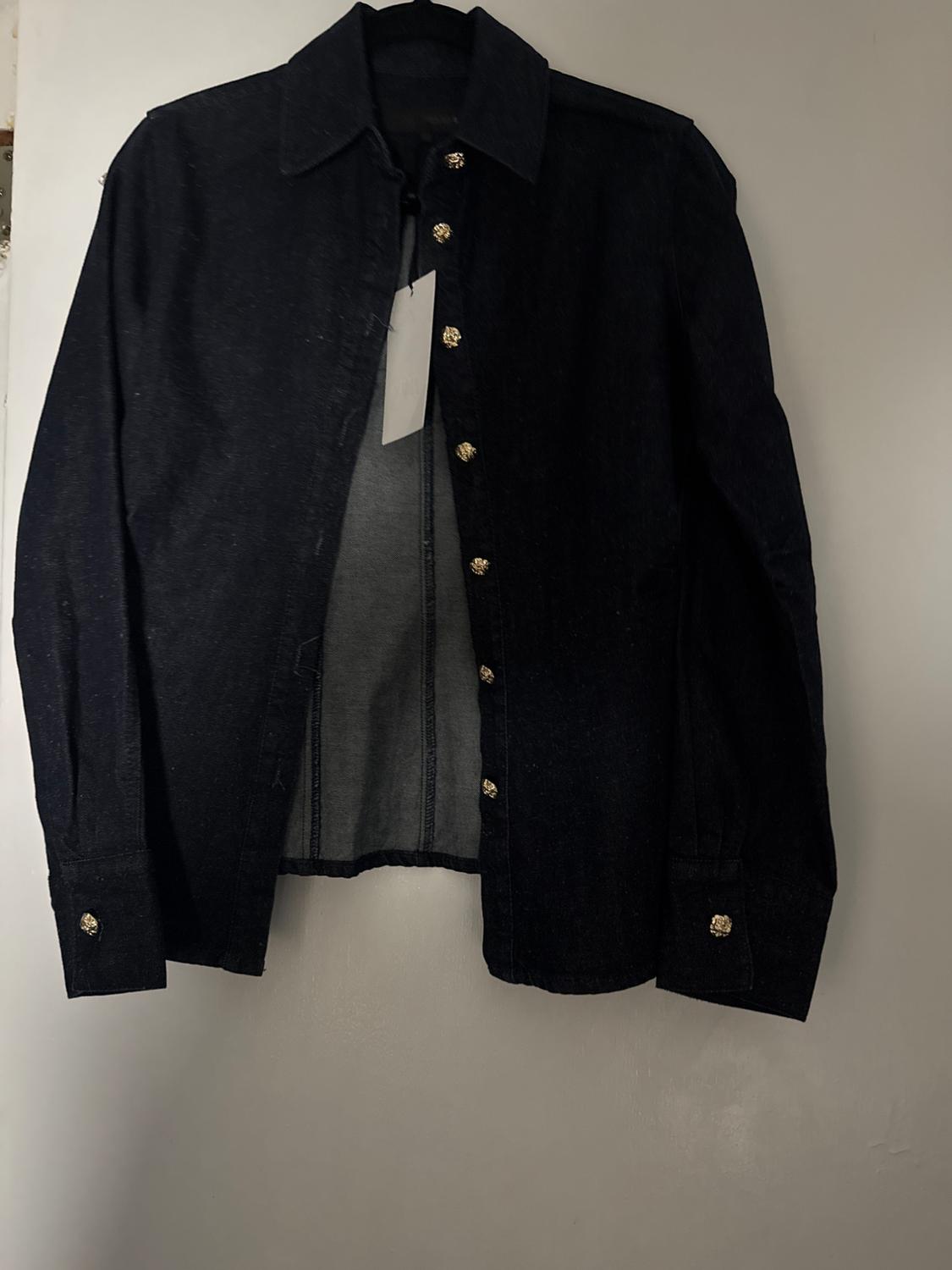 Black denim shirt hanging on a hanger, with gold buttons and a visible white tag in the center. White background.