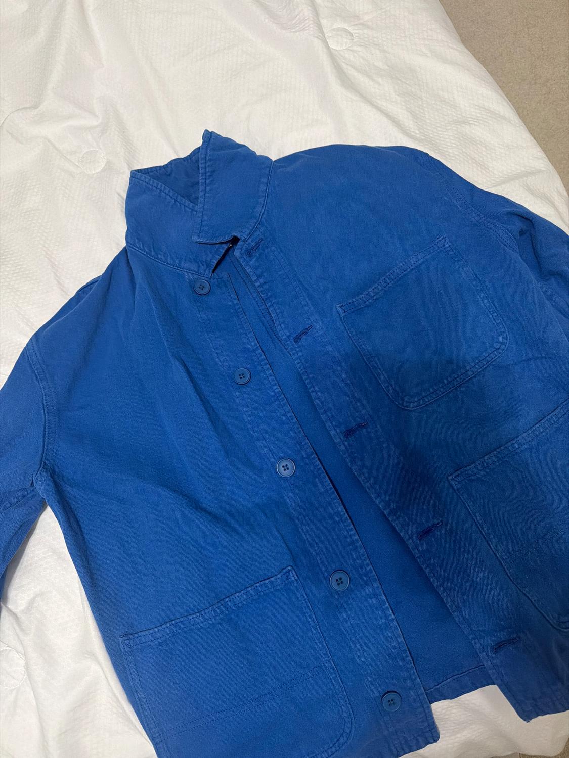 Blue jacket with buttons and three front pockets, placed on a bed with a white bedspread.