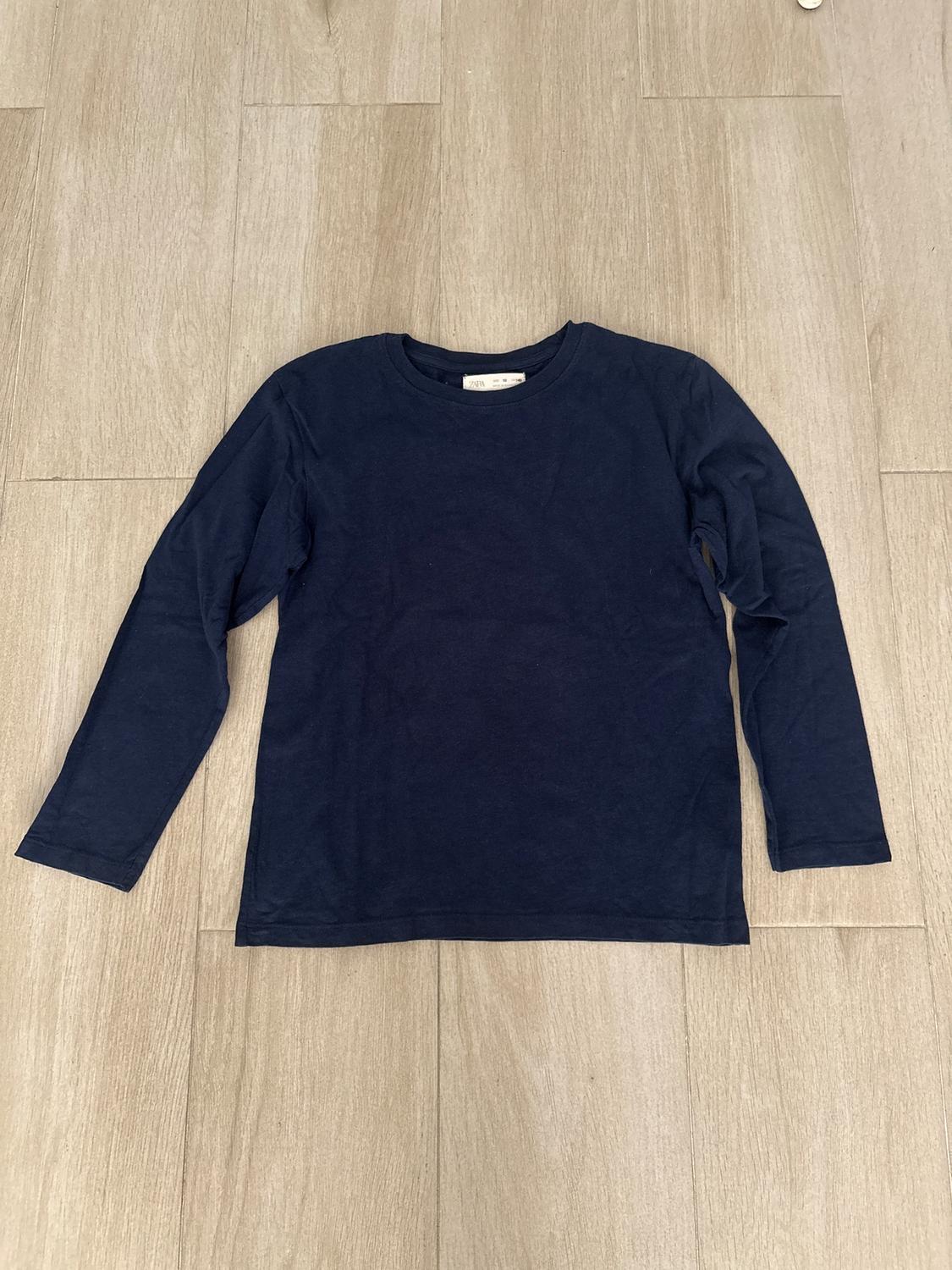 Navy long-sleeved T-shirt on a light wooden floor. Visible label on the collar.