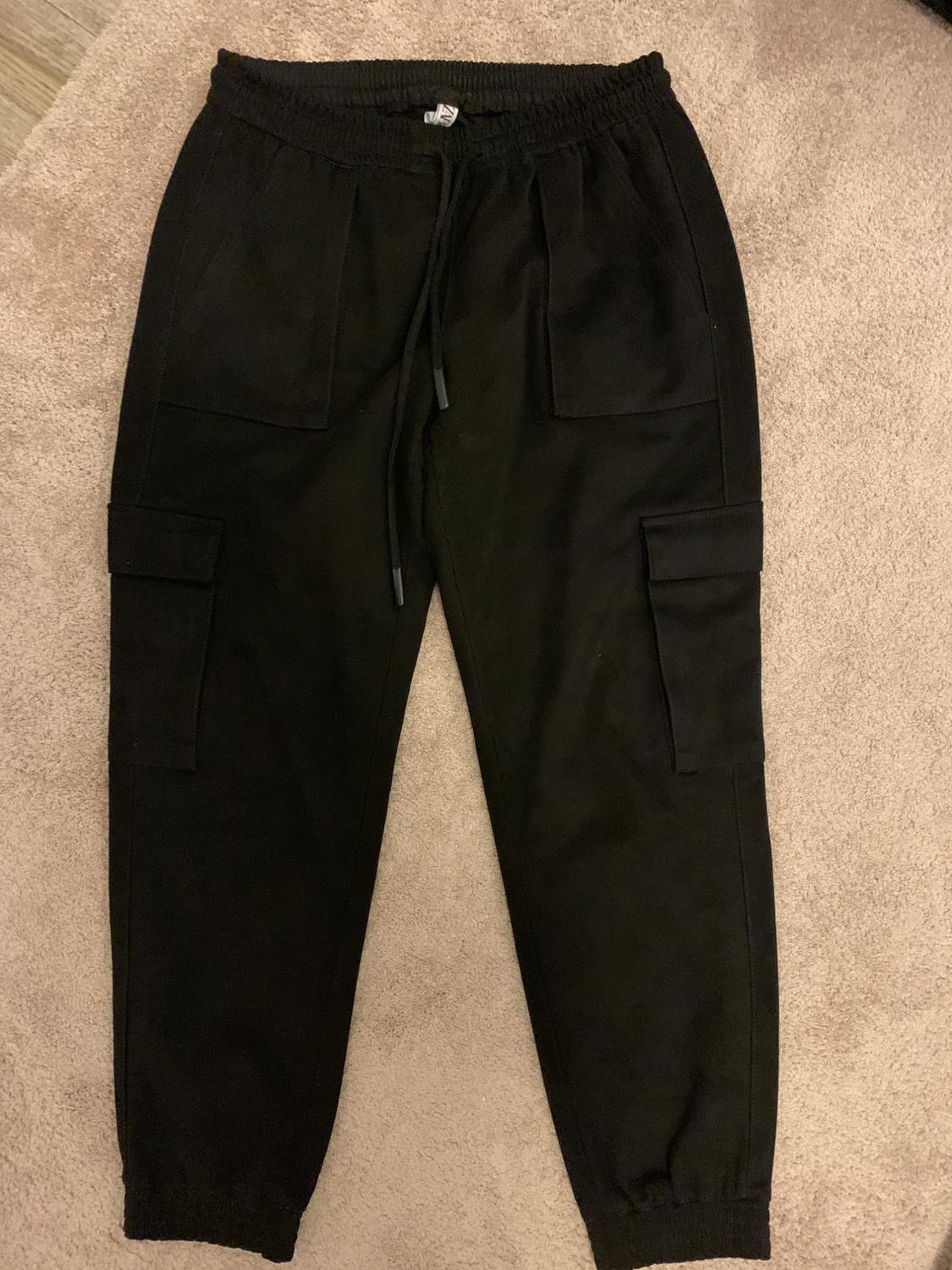 Black jogger-style trousers with side pockets and an elasticated drawstring waist, on a beige carpet.