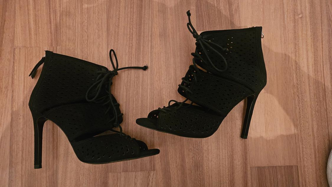 Pair of black high-heeled ankle boots with a perforated design, laces, and open toe, photographed from above on a wooden floor.
