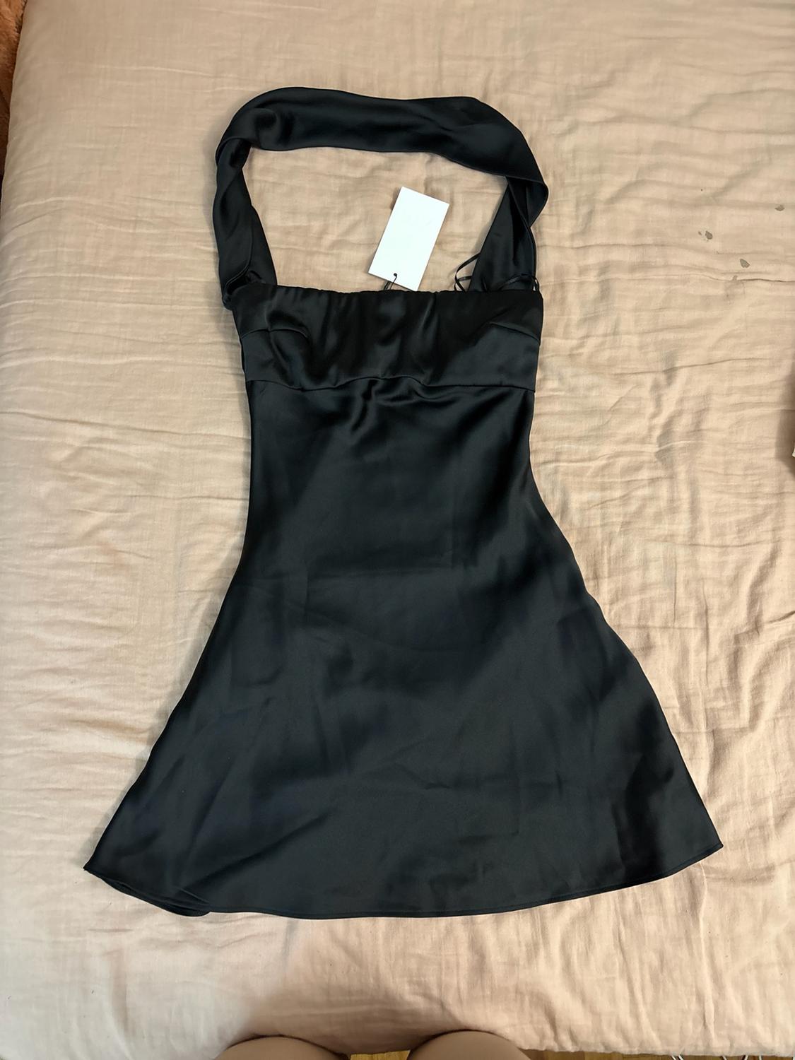 Short black satin dress with a halter neck and white label, casually laid out on a crumpled beige sheet.