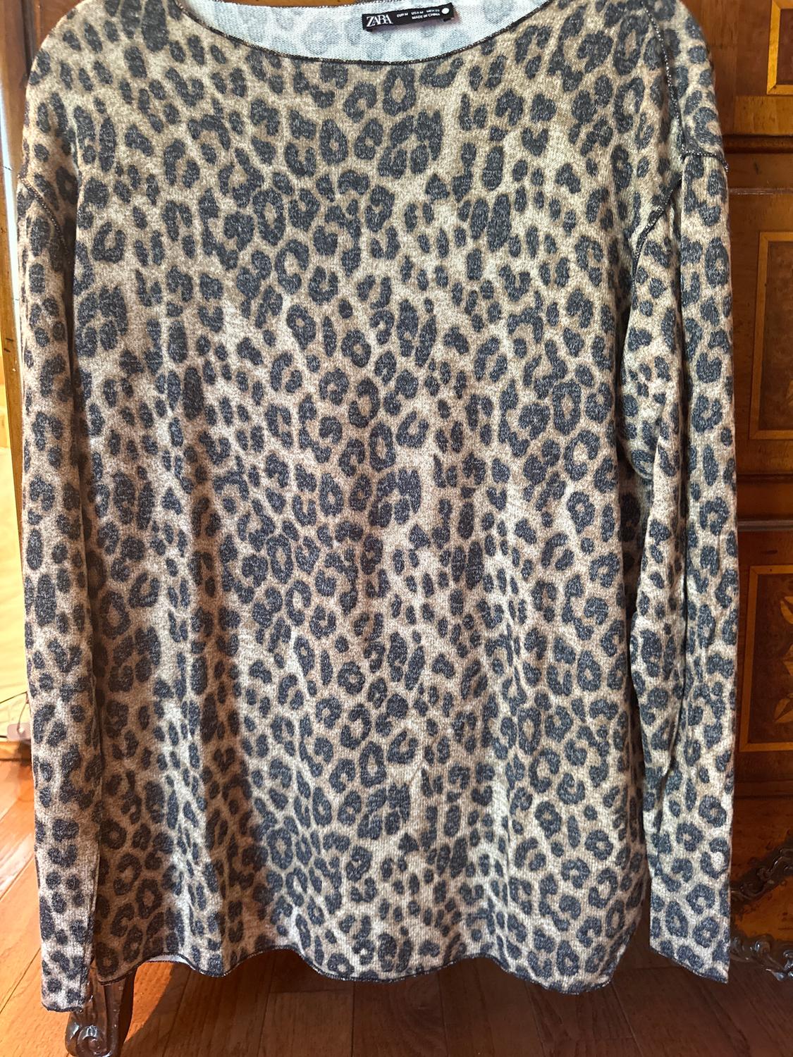 Long-sleeved leopard print jumper in brown and black tones, hanging in front of a wooden cabinet. Visible label on the collar.