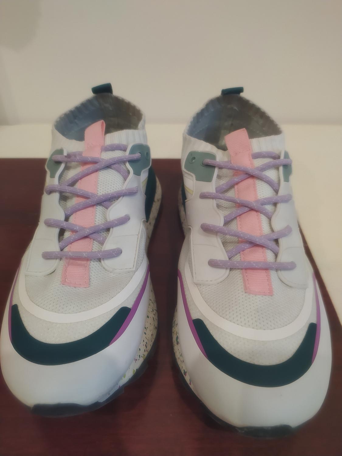 White trainers with pink and green details, lilac laces and a speckled sole, on a wooden background.