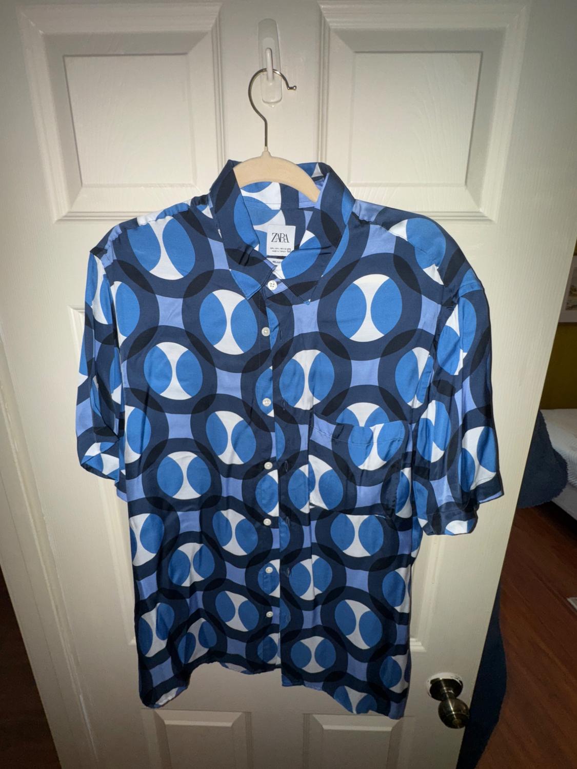 Blue and white geometric print short-sleeved shirt hanging on a white door. Visible tag: Zara.