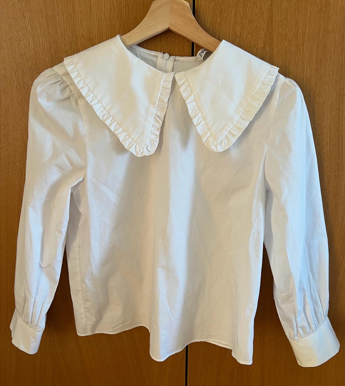 Long-sleeved white blouse with a large frilled collar, hanging on a hanger against a wooden background. Visible text: ZAR.