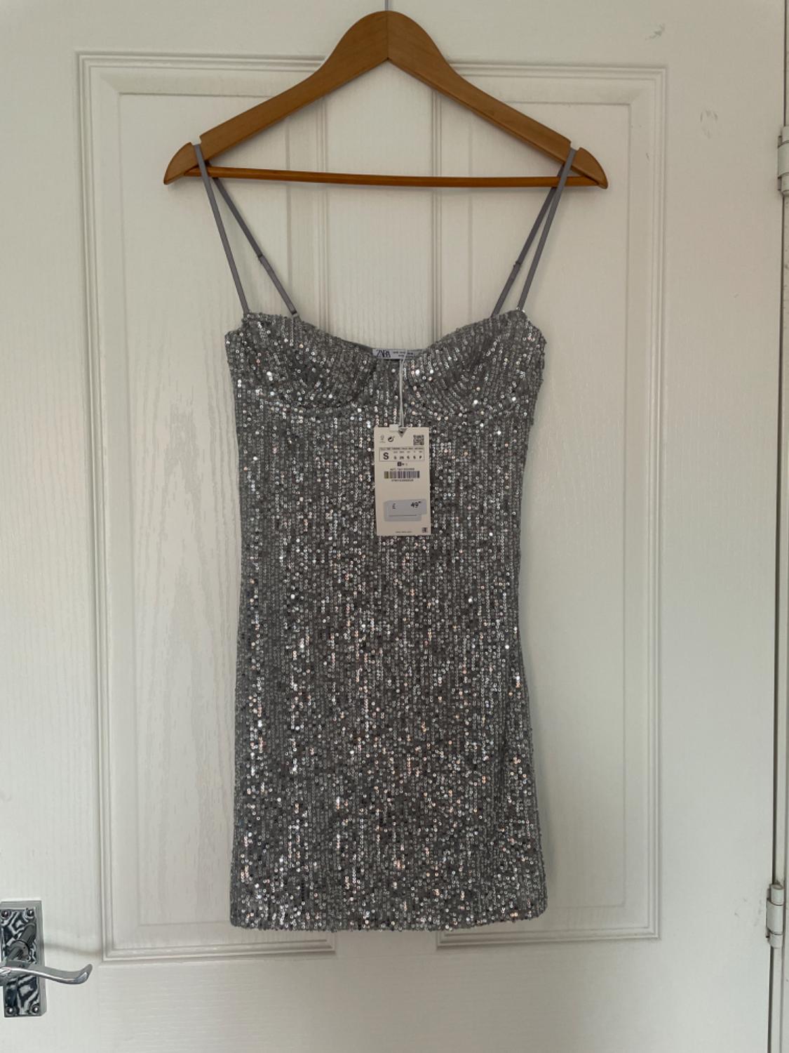 Silver sequinned mini dress with spaghetti straps, hanging on a hanger on a white door. Text: ZARA, S, £49.