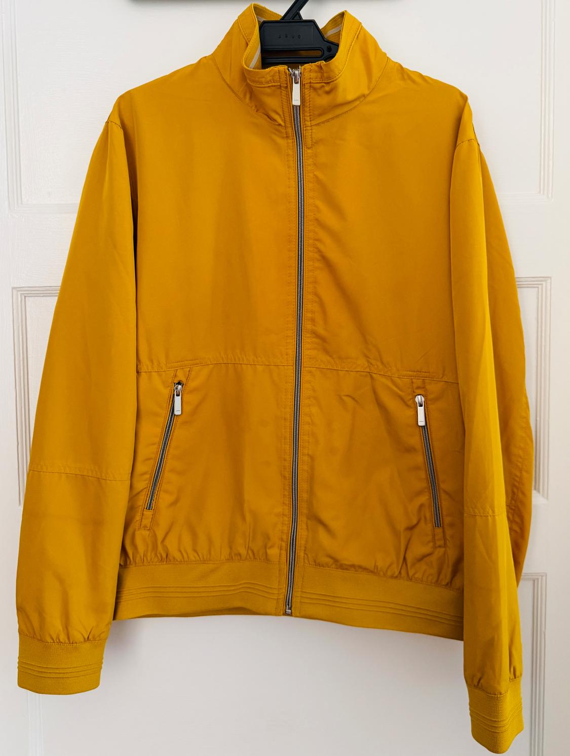 Mustard yellow casual jacket with zipper and pockets. It is hanging on a black hanger in front of a white door.