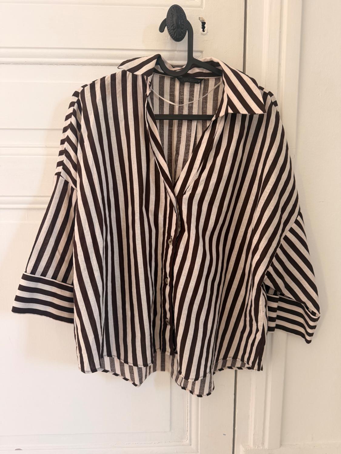 Loose-fitting shirt with dark and white stripes hanging on a white door. Wide sleeves with horizontal stripes on the cuffs.