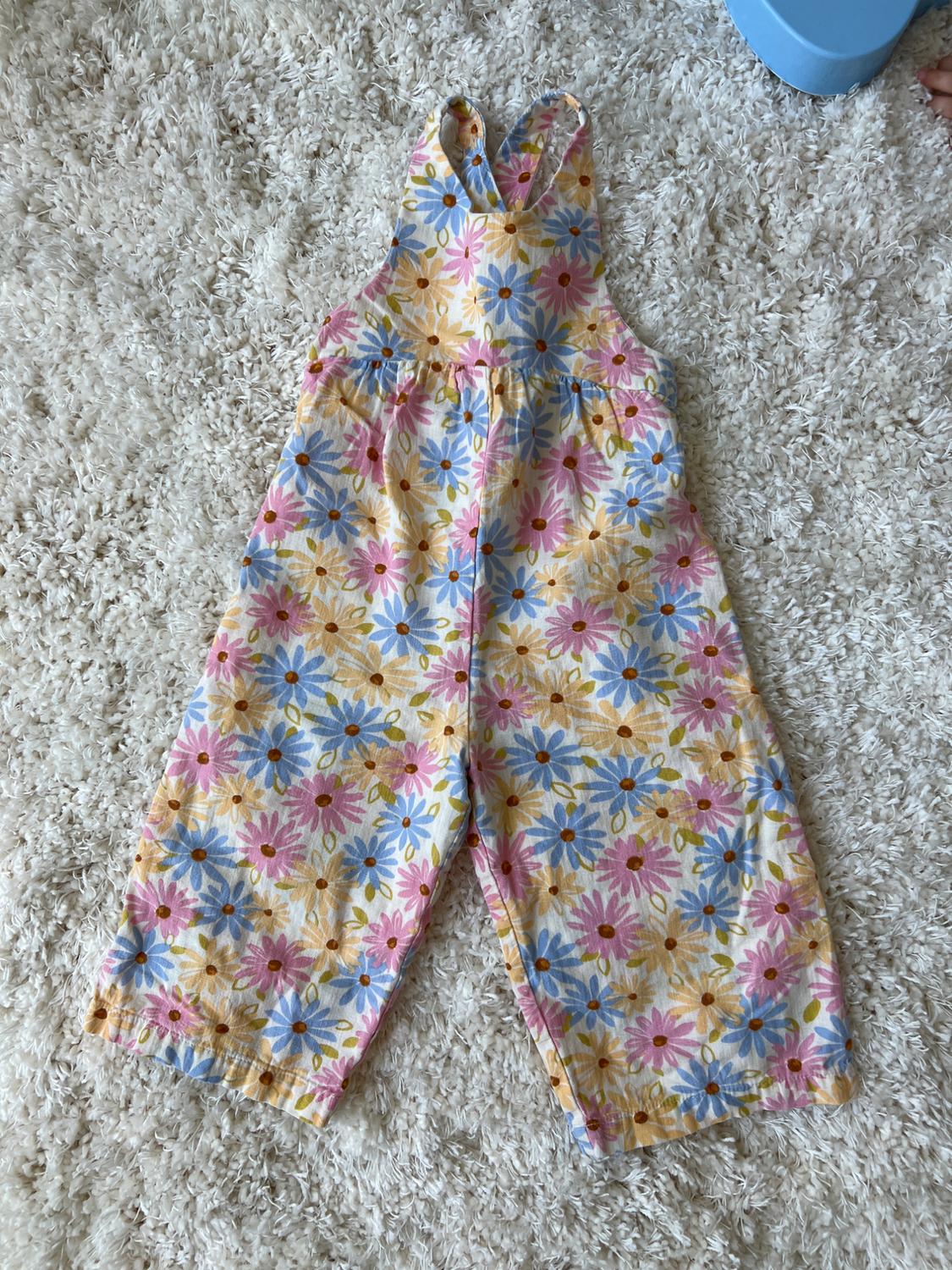 Kids' jumpsuit with a floral print in shades of pink, blue, and yellow on a white rug. Part of a blue object visible.