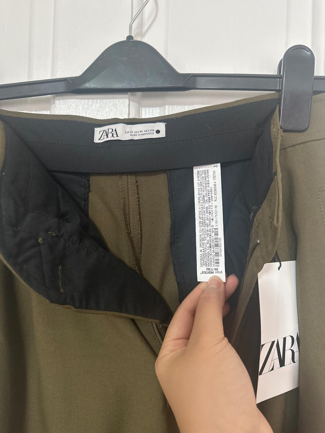 Olive green trousers hang from a black hanger, clearly displaying a Zara label. A hand holds the internal tag detailing the manufacturing.
