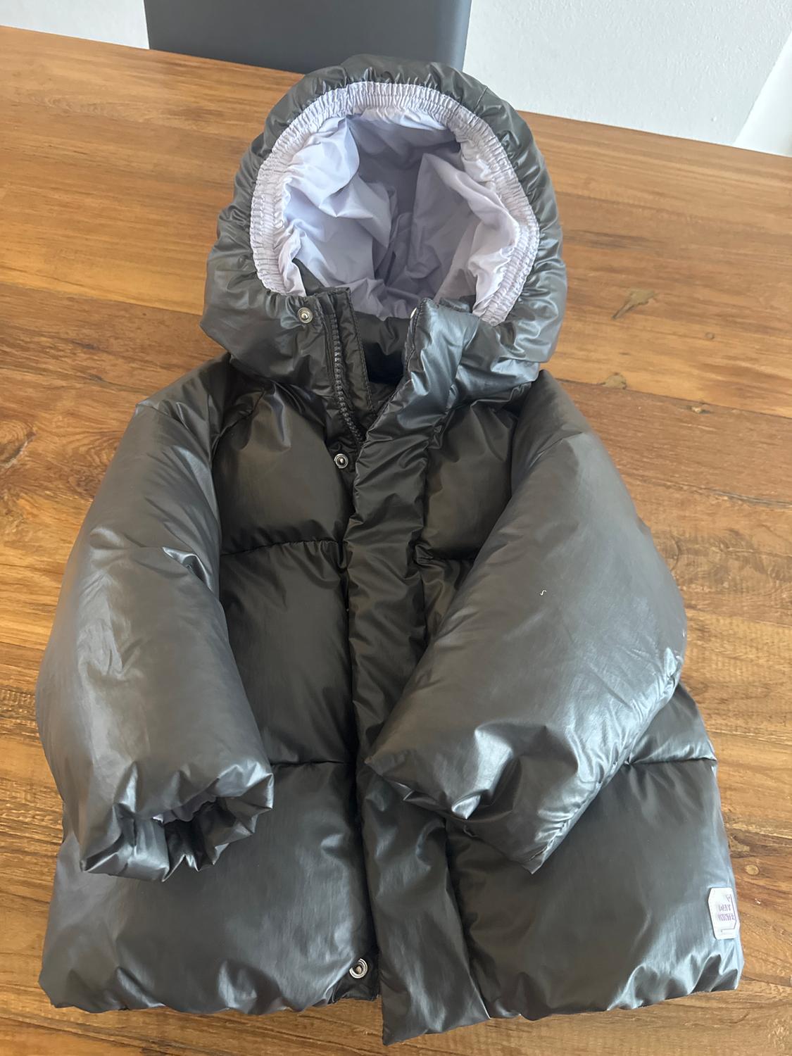 Grey padded jacket with white hood on a wooden table. The label reads "Baby Moncler".