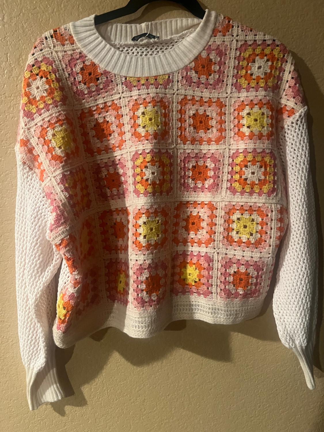 Knitted jumper hanging up, with a checked design in shades of pink, orange and yellow on a white background, on a beige wall.