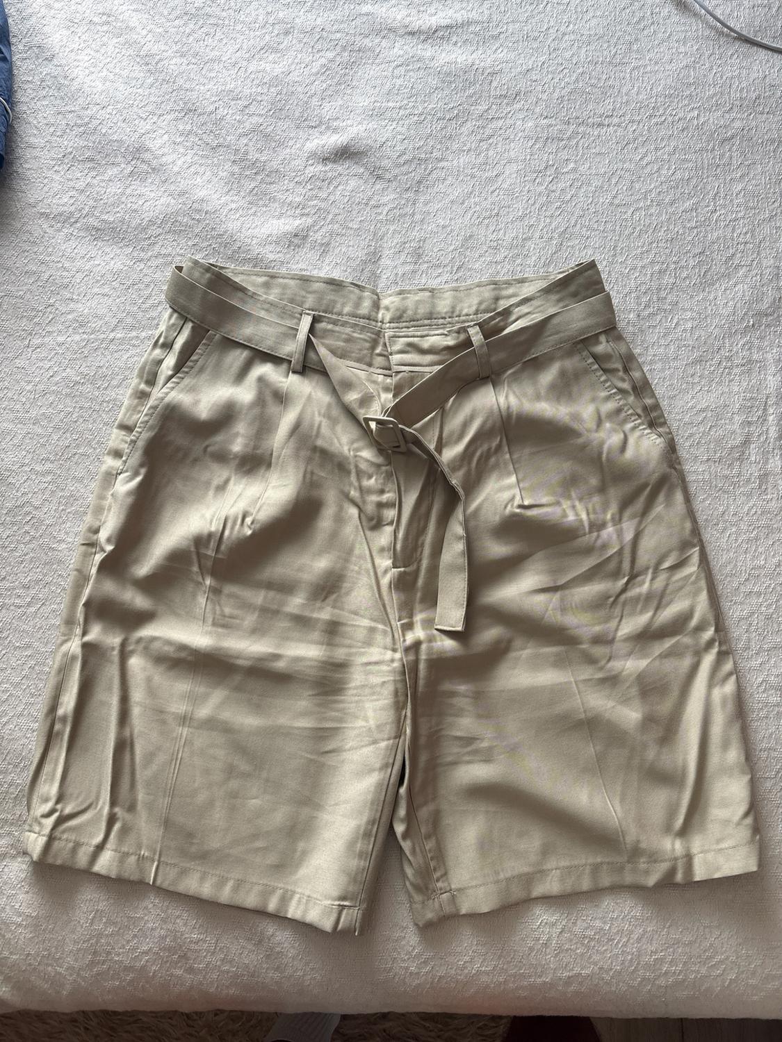 Beige pleated shorts with a matching belt on textured white fabric. Casual style overhead composition.