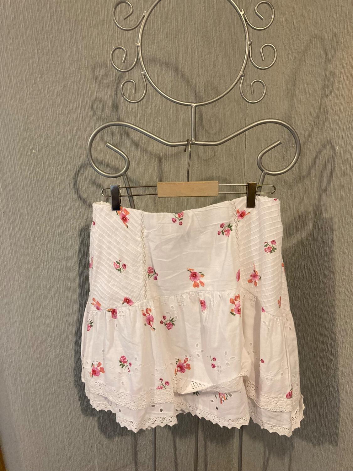 White short skirt with pink floral print and openwork details, hanging on a decorative metal hanger against a grey wall.