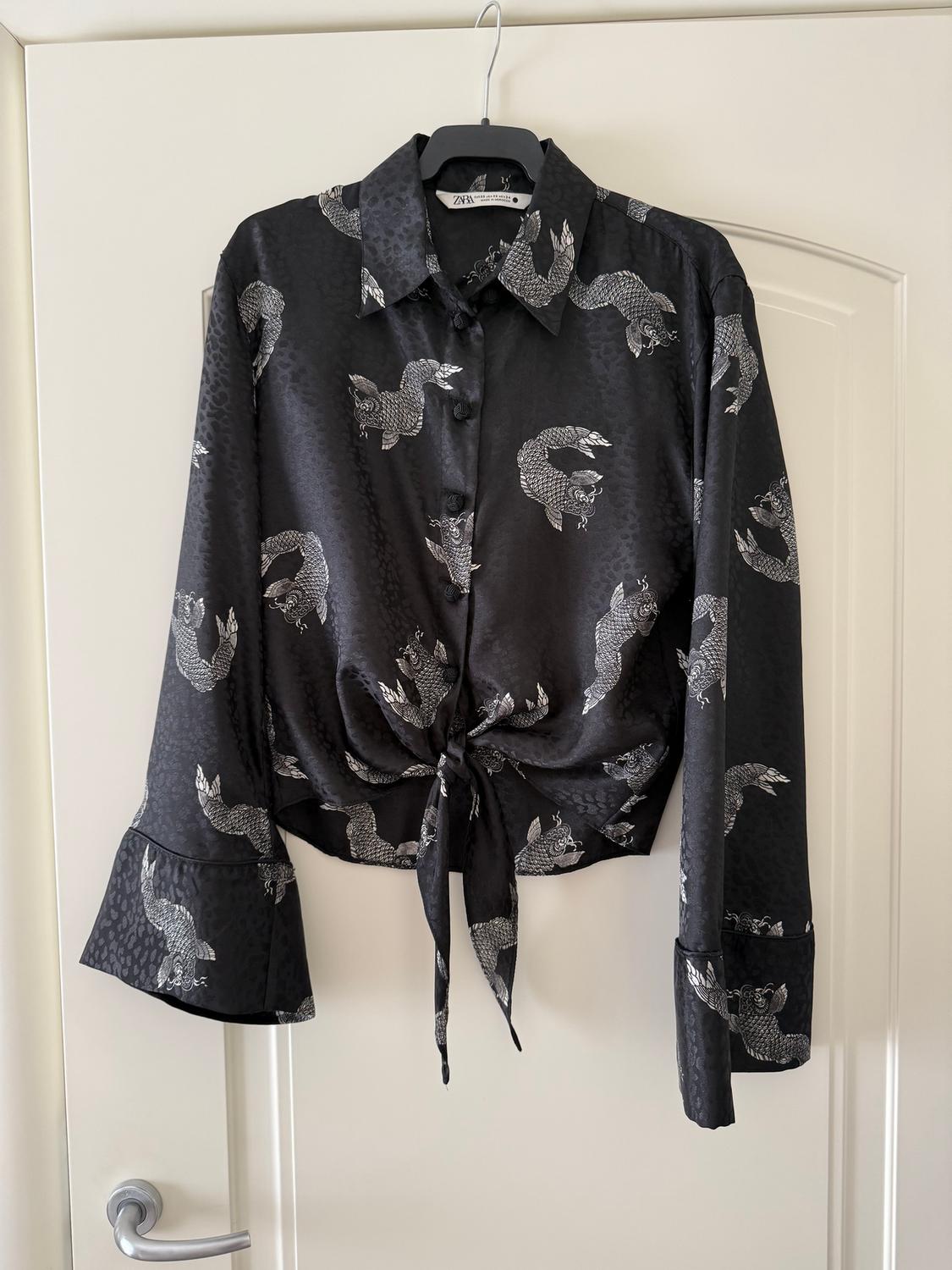 Black blouse with a white dragon print, long flared sleeves, and a front knot, hanging on a hanger over a white door.