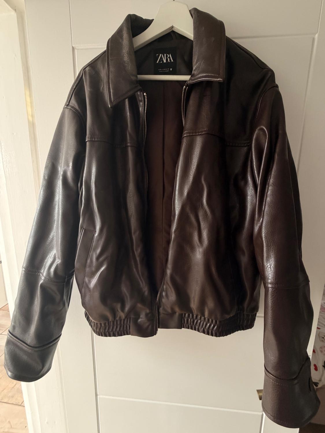 Dark brown jacket with a zip hanging on a white hanger over a door. Inner label with the text ZARA.