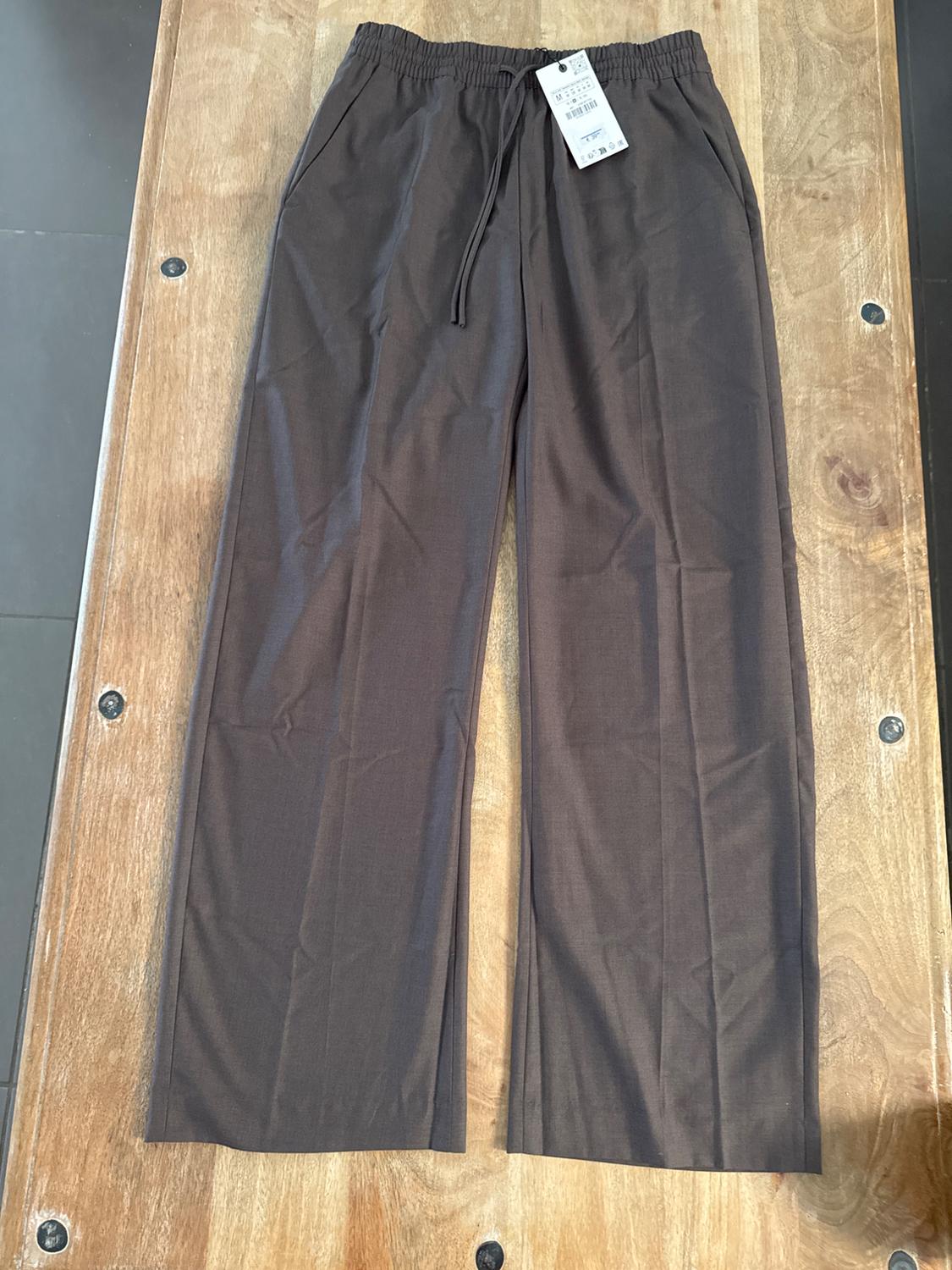 Brown wide-leg trousers with elasticated waistband and drawstring, tagged, on a wooden table.