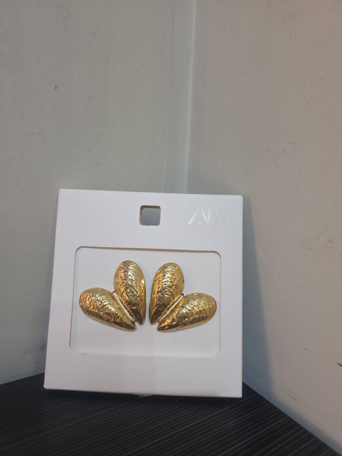 Gold-tone shell design earrings on a white stand with the text ZARA, on a dark surface in front of a white wall.