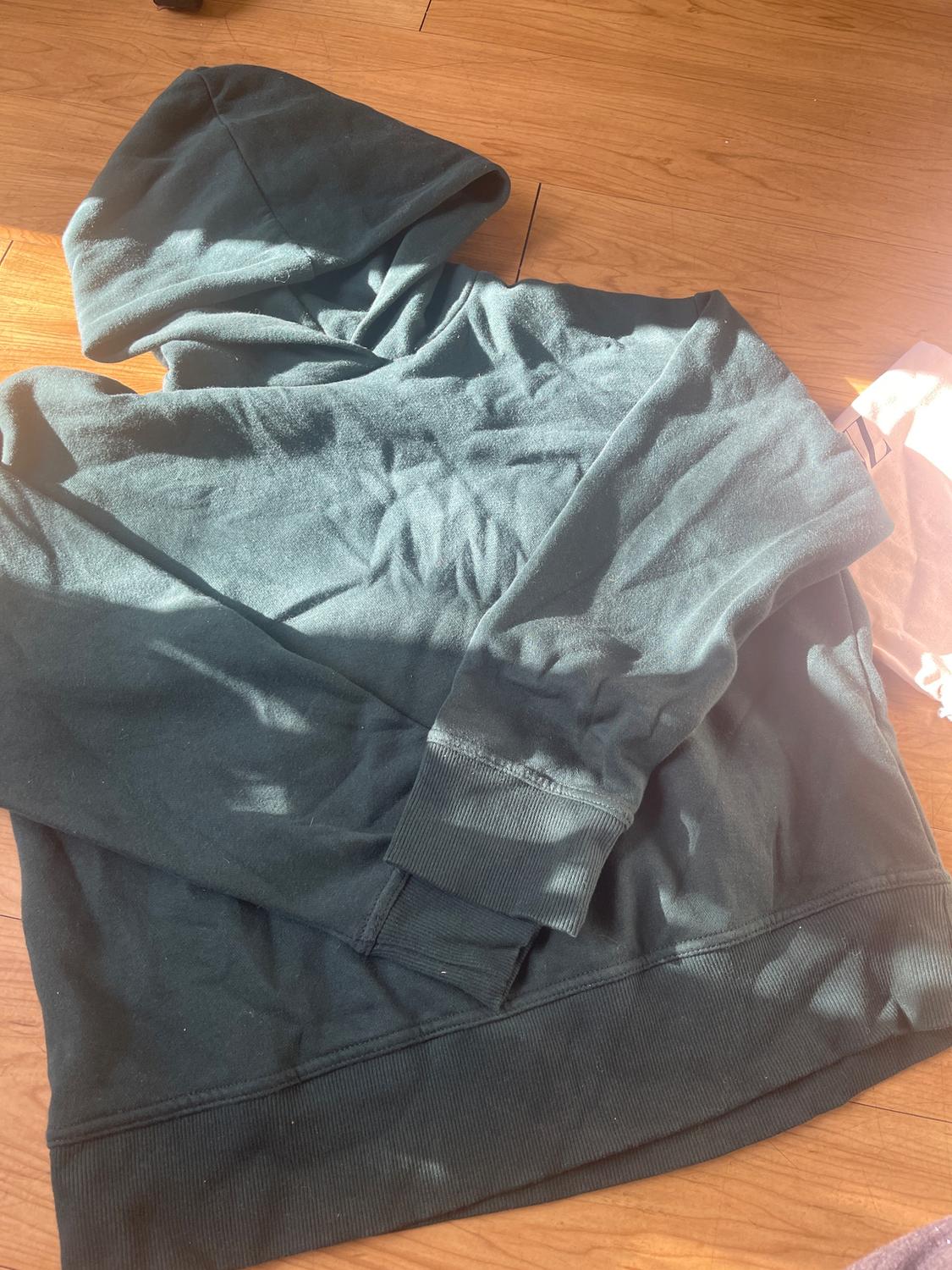 Dark green hoodie on a wooden floor, illuminated by natural light. Wrinkled fabric and soft texture visible.