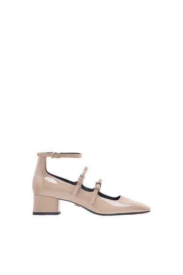 Beige patent leather shoe with a low block heel, three-strap design with buckles, viewed in profile against a white background.