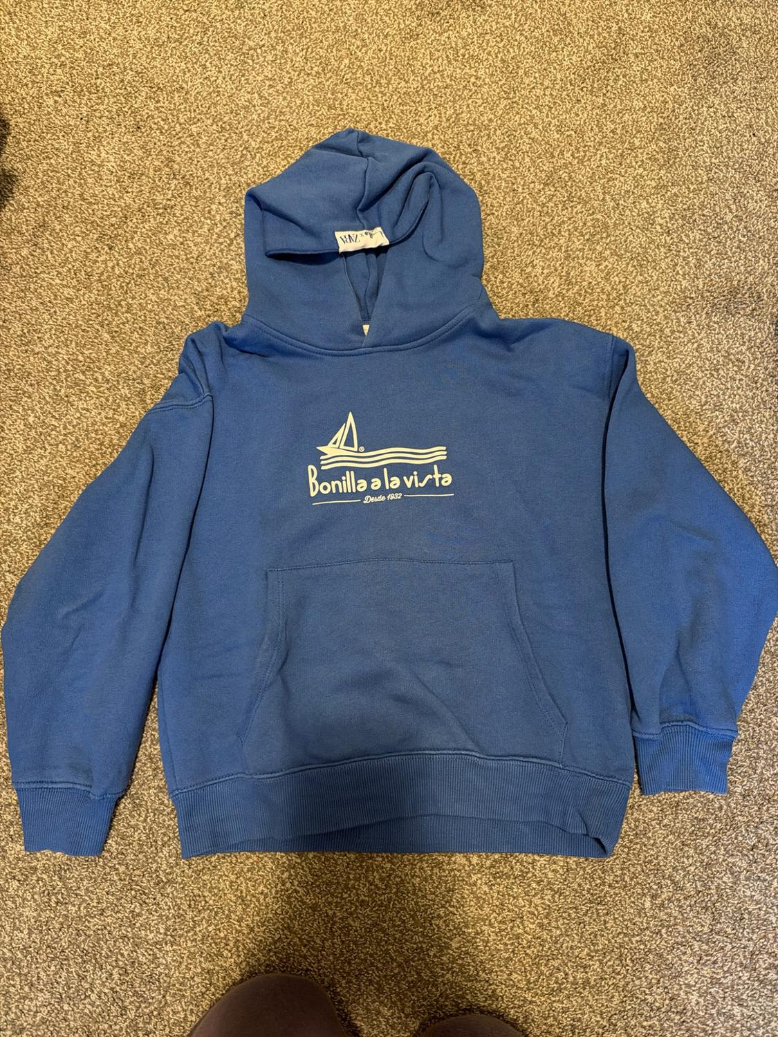 Blue hoodie on a rug. Includes boat graphic and the texts "zara", "Bonilla a la vista" and "- Desde 1932 -".