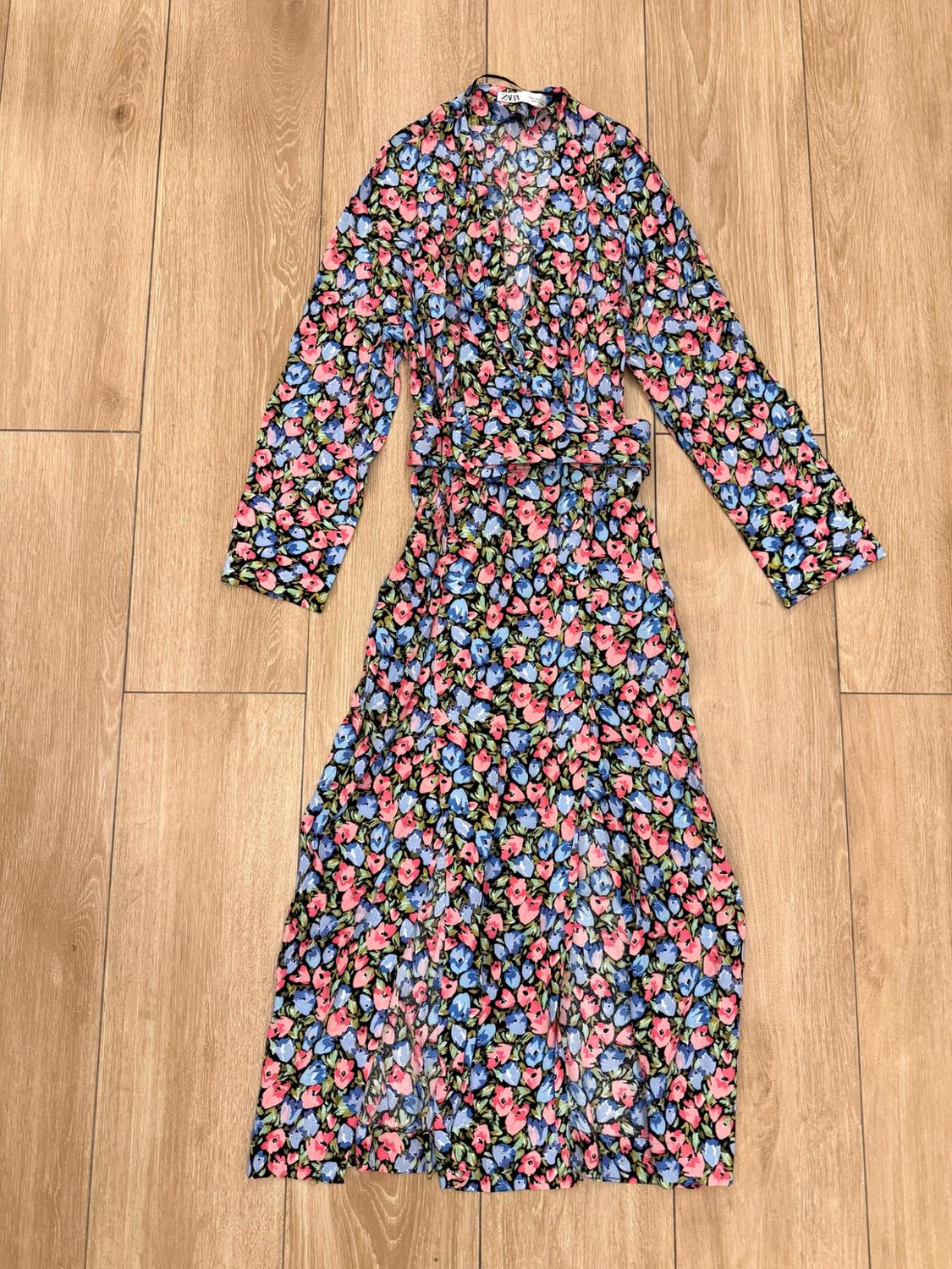 Wrap dress with a pink and blue floral print on a black background, long sleeves, a belt and a ZARA label, on a wooden floor.