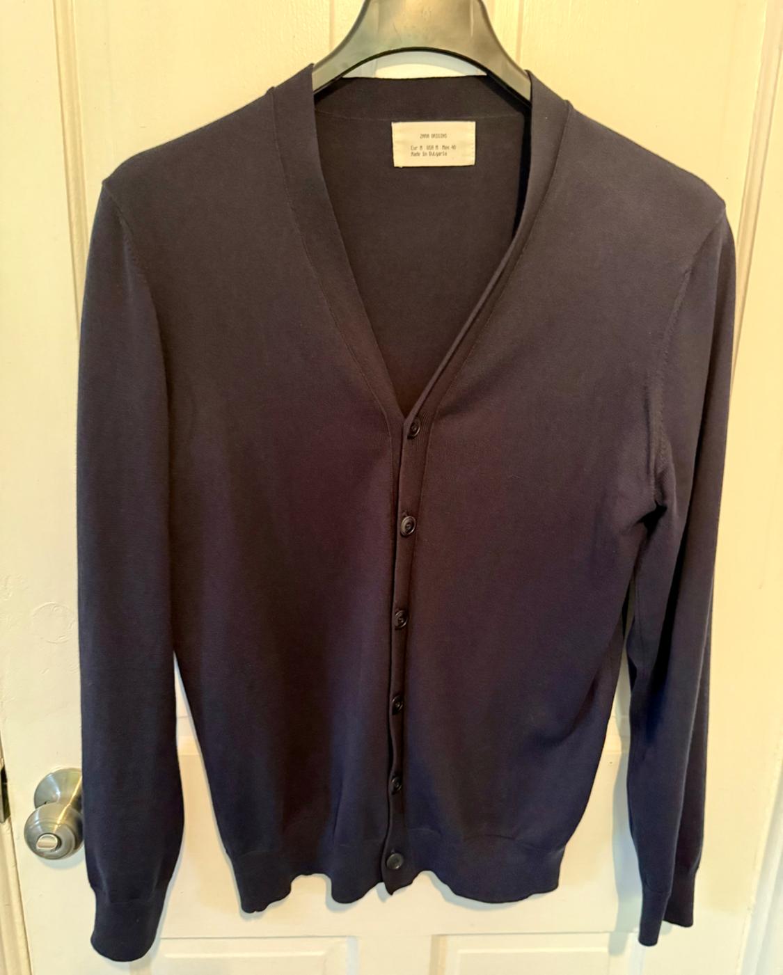 Black, long-sleeved cardigan hanging on a clothes hanger, with buttons down the front and a visible label on the neck, over a white door.