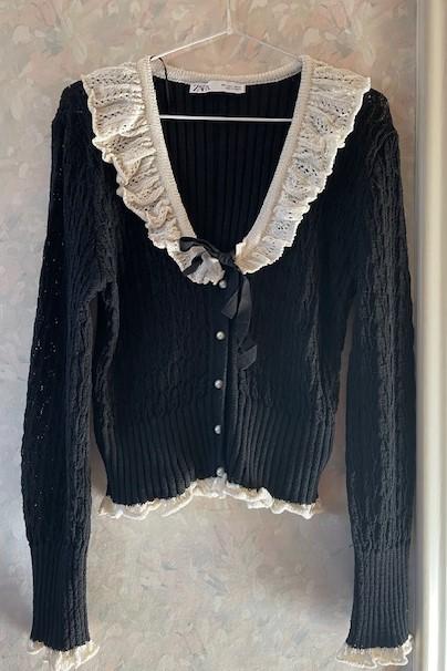 Black knitted cardigan with white ruffles on the collar and cuffs, hanging on a hanger against a light background.