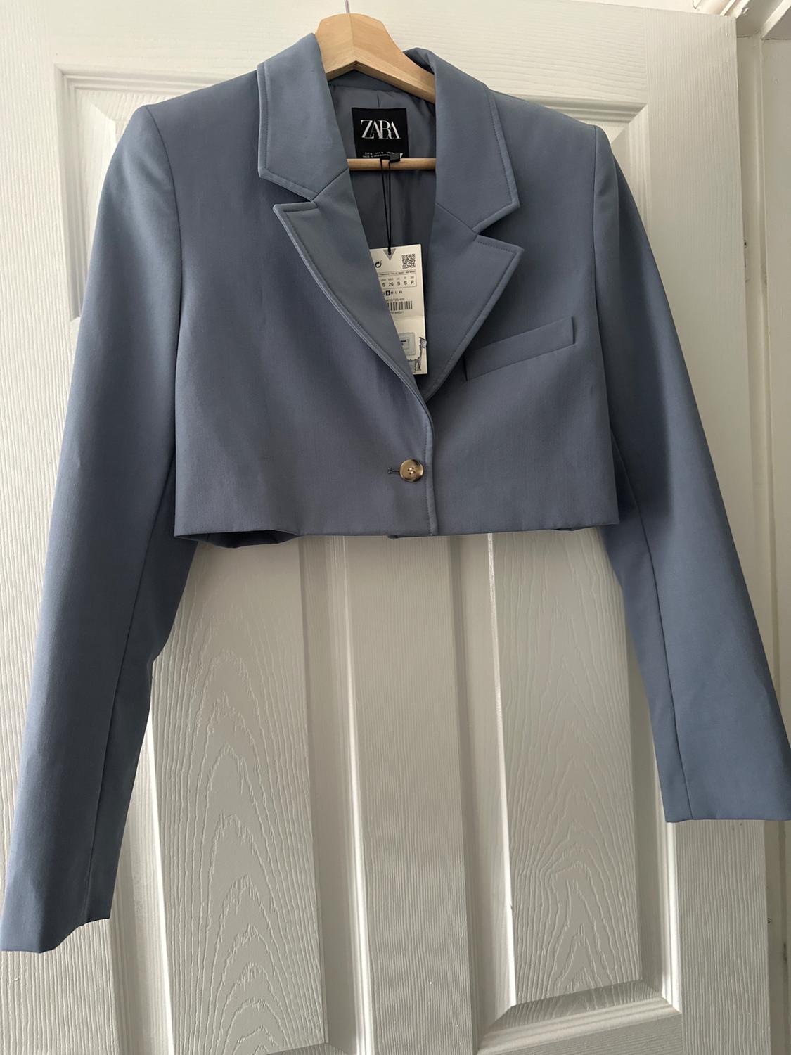Short blue jacket hung on a white door, with visible label and "ZARA" brand on the inside of the neck.