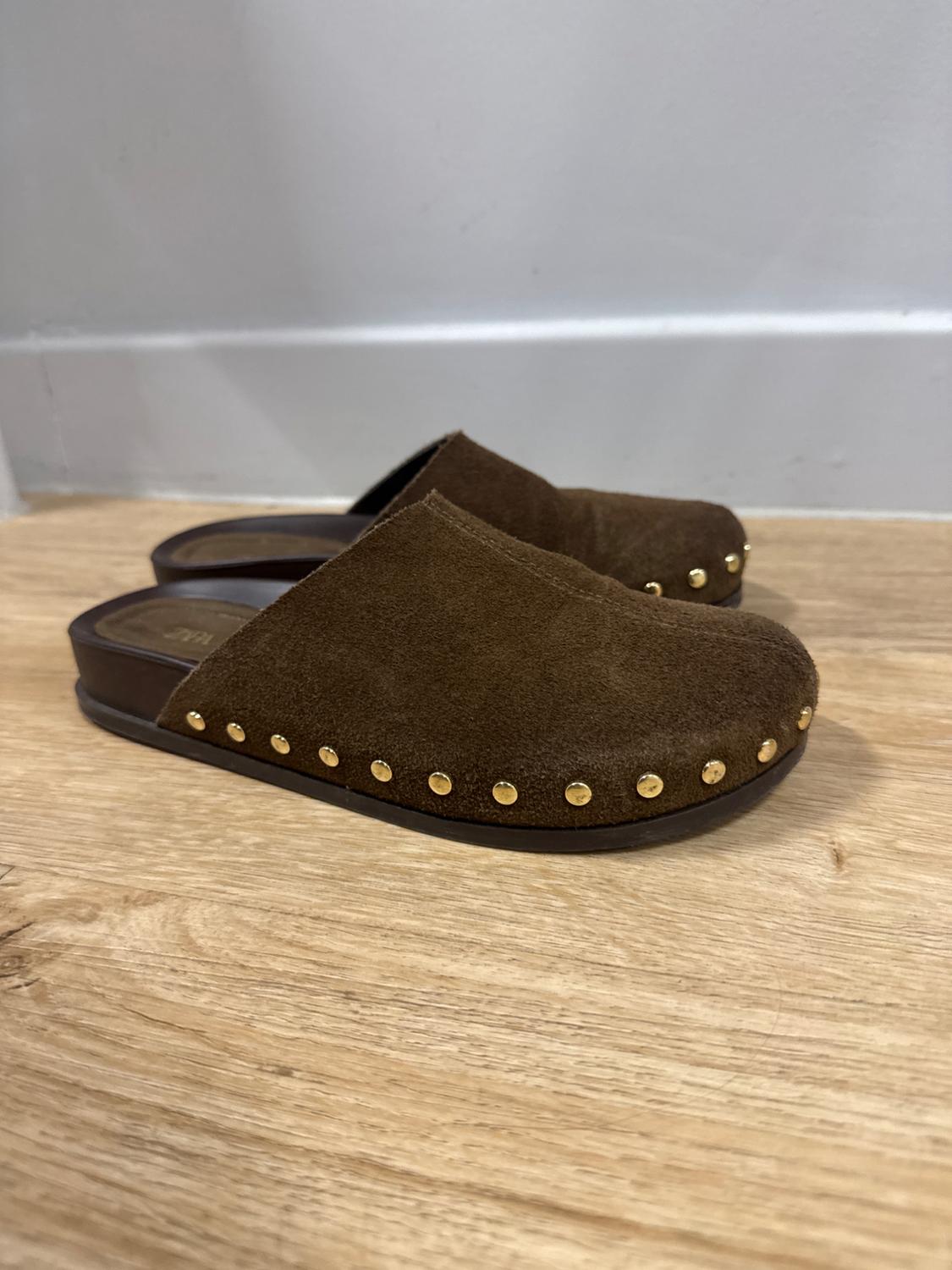 Brown suede clogs with a dark sole and gold studs on a light wood floor in a relaxed setting. Text: ZARA.