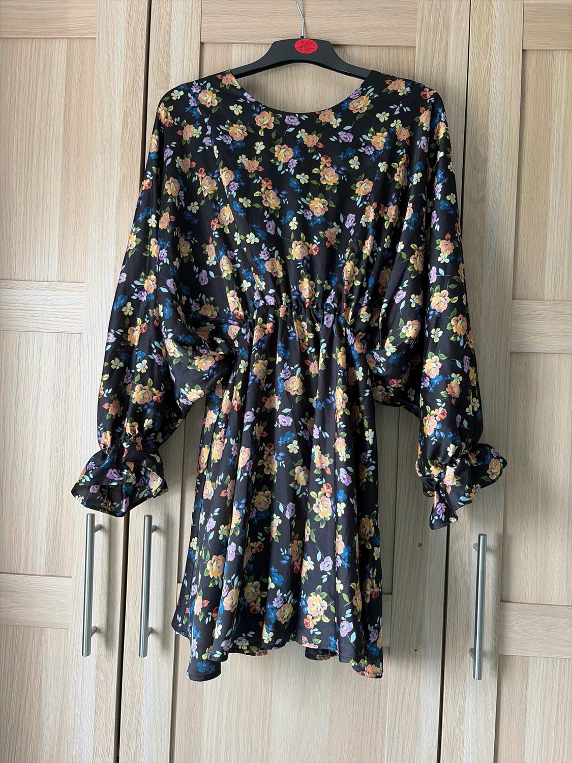 Short black dress with floral print, long sleeves and fitted waist, hanging on a hanger in front of a wooden wardrobe.